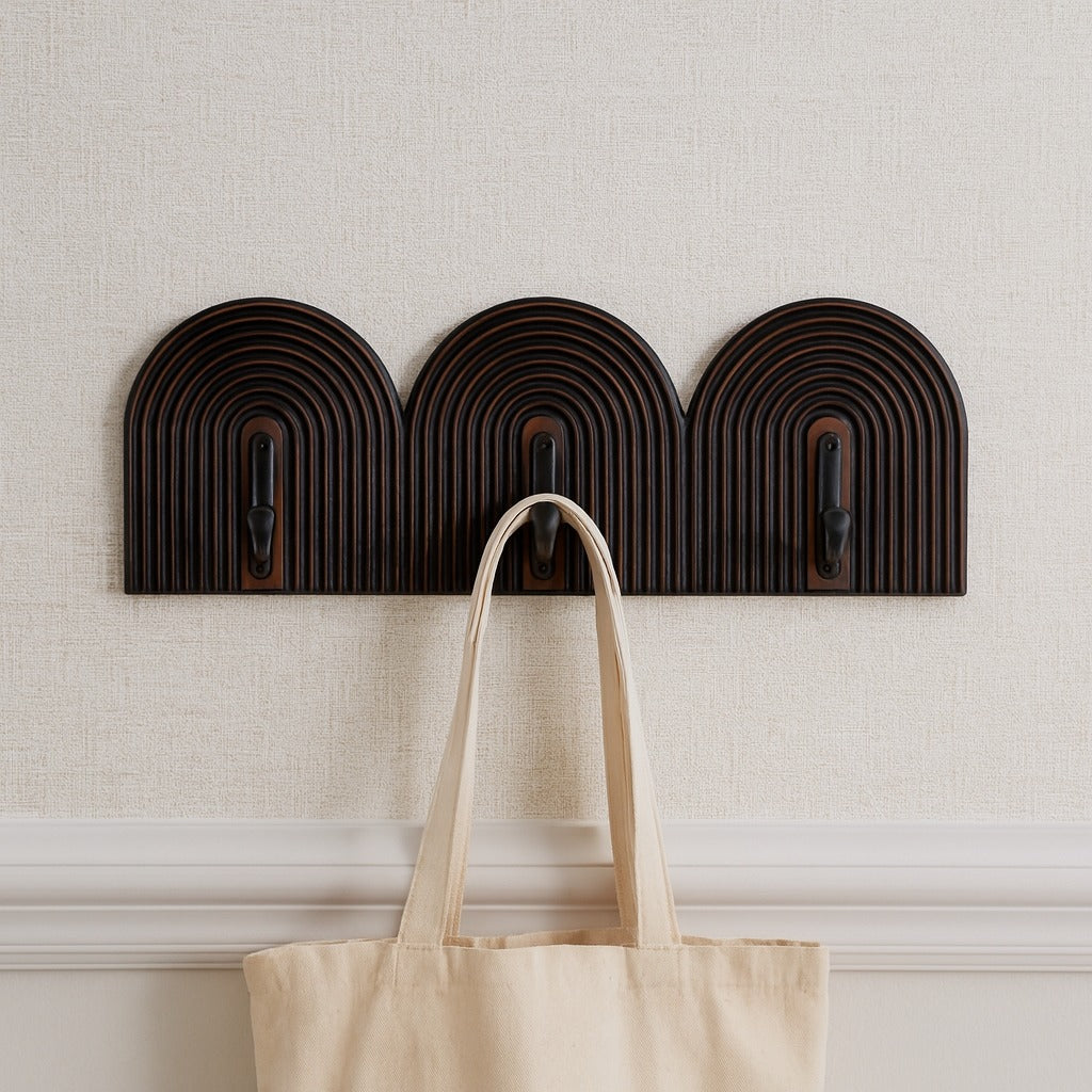 Three dark brown and black arch wall hooks with a beige tote bag hanging on one of them against a light gray wall.