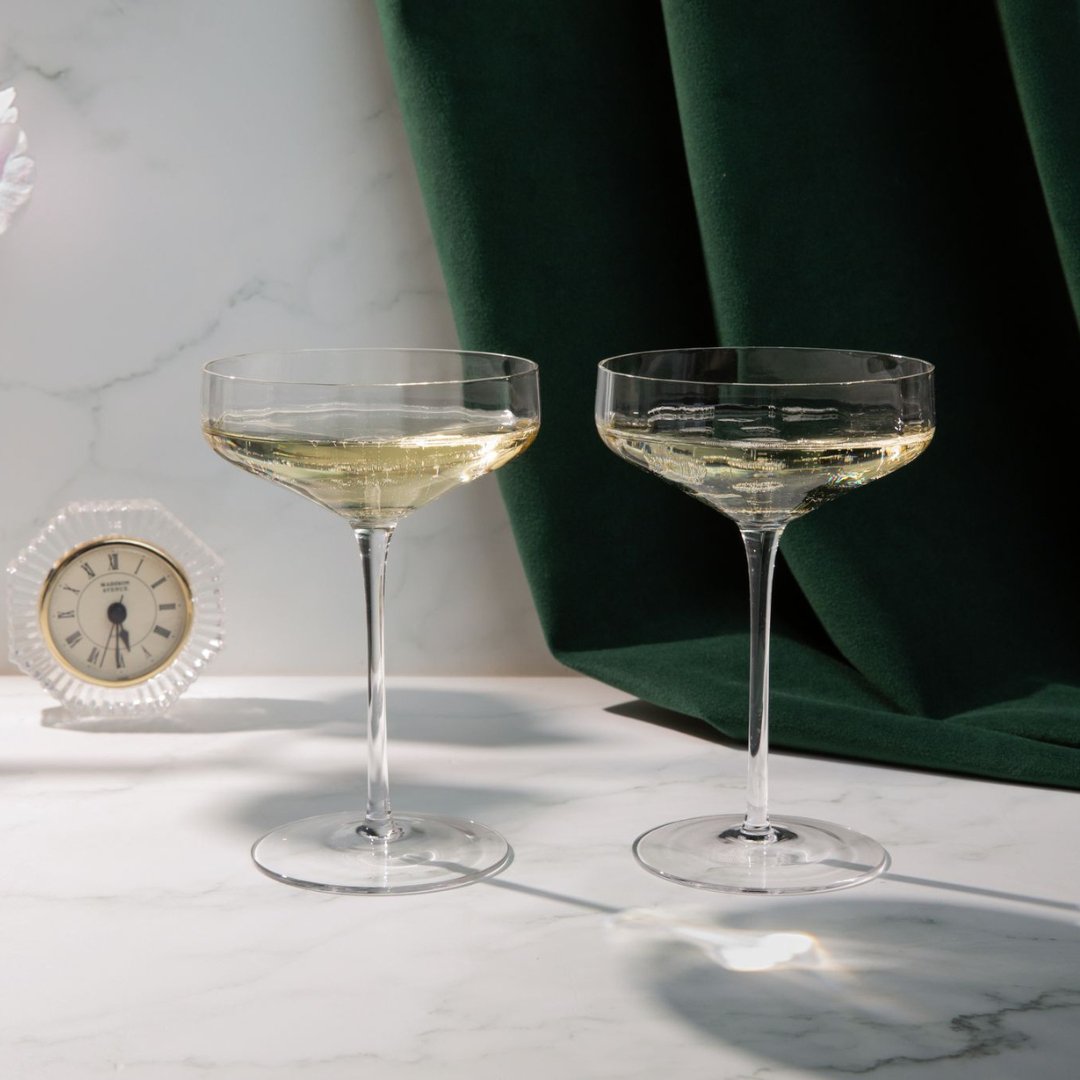 Two champagne Angled Coupe Cocktail Glasses with a small clock on a marble surface